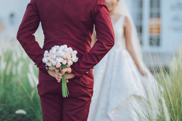 Close up shot of the groom hides a bouquet of flowers behind to bring to his beautiful Asian bride, blurred at woman.