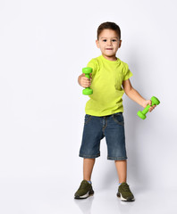 Little boy in yellow t-shirt, denim shorts, khaki sneakers. Smiling, holding green dumbbells, posing isolated on white background