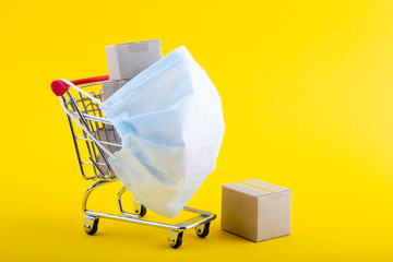 Trolley with medical mask and cardboard boxes on a yellow background. Online shopping during epidemic and quarantine. Protect Yourself medical mask. The concept of safe shopping and delivery.