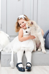 Beautiful little curly-haired girl in a white dress with a young dog of a husky breed, shot in bright studio scenery.