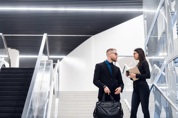 Business couple on a stairs talking and smiling. Man wearing suit and glasses with a bag talking with a woman holding tablet computer.