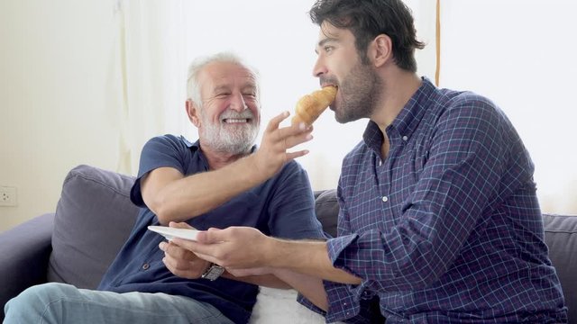 Happy family with senior father and son eating bread with delicious together with cheerful on sofa, young man conversation with elderly on couch at living room, lifestyle and relation concept.