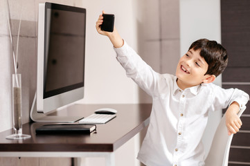 A boy standing in front of a computer taking a selfie with smartphone