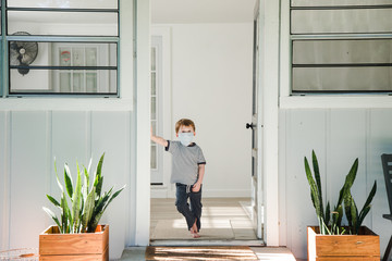 Young Caucasian Boy Standing in Doorway of a House Wearing a Medical Mask Looking at Camera