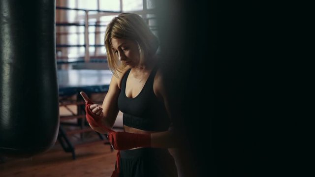 Young beautiful woman wrapping hands with red boxing wraps in the gym near the ring.