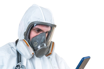 a doctor in protective medical coveralls and full face respiratory mask looks at his mobile phone in hand isolated on a white background