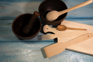 On a wooden table are two bowls of red clay. On a cutting board are wooden cutlery - small and large spoons, a fork and a spatula.