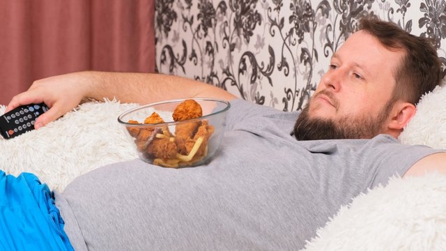fat man with a big belly is lying on the couch with fast food and is bored in front of the TV with the remote control in his hand, close-up.