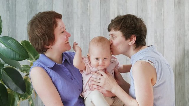Two young smiling mothers and a baby girl. Moms hold the baby in their arms, hug and kiss. Home interior. Homosexual lesbian family.