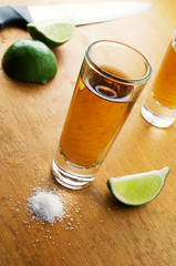 A tequila filled shot glass over a wooden tabletop with a slice of lemon  and a pinch of salt to...