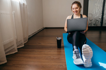 A sporty woman in sportswear is sitting on the floor with dumbbells and a protein shake or a bottle...