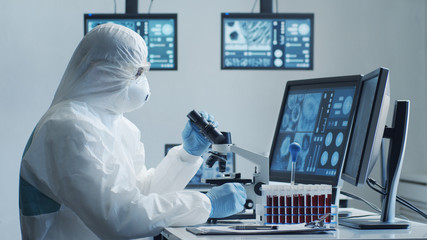 Scientist in protection suit and masks working in research lab using laboratory equipment: microscopes, test tubes. Coronavirus 2019-ncov hazard, pharmaceutical discovery, bacteriology and virology.