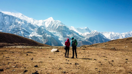 Fototapeta premium A couple walking on the Annapurna Circuit Trek, Himalayas, Nepal. Snow caped Annapurna chain in the back. Clear weather, dry grass, snowy peaks. High altitude, massive mountains. Freedom and adventure