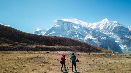Fototapeta premium A couple walking on the Annapurna Circuit Trek, Himalayas, Nepal. Snow caped Annapurna chain in the back. Clear weather, dry grass, snowy peaks. High altitude, massive mountains. Freedom and adventure