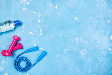 Pink dumbbells, jump rope, bottle of water on blue background. Sport, training, slimming, healthy lifestyle, fitness, home workout concept. Sport equipment. Copy space