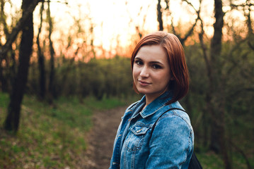 Portrait of young and beautiful redhead woman with big brown eyes in blue jean jacket on sunset. Horizontal shot - Girl (female model) with soft skin and pretty smile posing to camera in forest road.