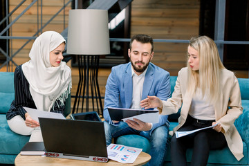 Naklejka premium Handsome bearded Caucasian businessman and two pretty multiracial businesswomen consulting each other, working together on joint financial project in the modern loft office room