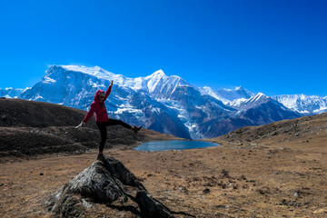 A woman wearing pink jacked and black leggings, standing on a rock in the nearby the Ice Lake, Annapurna Circuit Trek in Himalayas, Nepal. Snow caped Annapurna Chain in the back. Playful time. Freedom
