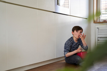 Portrait of boy sitting on kitchen floor with red mixing bowl and egg beater tasting batter