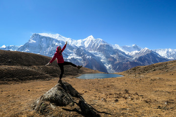 A woman wearing pink jacked and black leggings, standing on a rock in the nearby the Ice Lake, Annapurna Circuit Trek in Himalayas, Nepal. Snow caped Annapurna Chain in the back. Playful time. Freedom