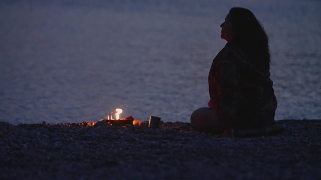 A woman sits by a campfire on the beach on an autumn evening