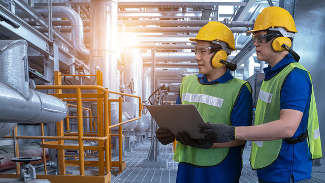 Industrial Engineer In Hard Hat Uses Tablet While Standing In The Industry Manufacturing Factory. Oil And Gas Refinery Background. Two Factory Worker In A Hard Hat On Heavy Equipment And Pipeline