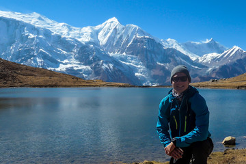 Fototapeta na wymiar A man in hiking outfit squatting at the shore of Ice Lake, Annapurna Circuit Trek, Himalayas, Nepal. He is enjoying the idyllic landscape. High, snow caped Annapurna chain in the back. Achievement