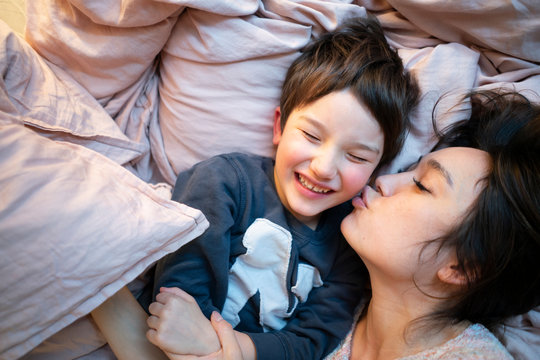 Portrait of laughing little boy lying  on bed at home kissed by his mother
