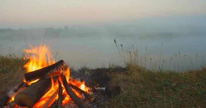 Fire by the river. Bonfire on the river bank on an early foggy morning. A bonfire burns on the high sandy bank of the river. Red fire bonfire near the water. Camping vacation for tourists or fishermen