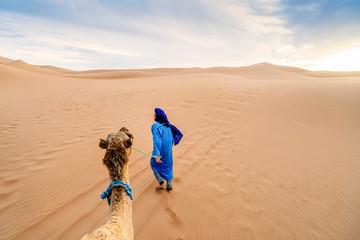 A Bedouin man wearing blue walking with camel through yellow sand dunes of Sahra Dessert