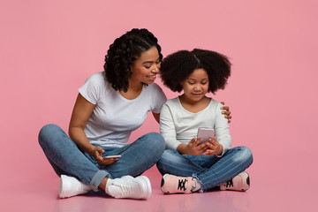 Naklejka na ściany i meble Parenting Control Apps. Little Black Girl Holding Smartphone Sitting Next To Mom