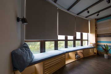 Motorized roller shades in the interior. Automatic roller blinds beige color on big glass windows. Remote Control Shades are above the windosill with pillows. Summer. Green trees outside. - obrazy, fototapety, plakaty
