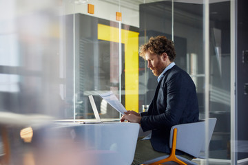 Businessman sitting at desk in office with laptop reading papers