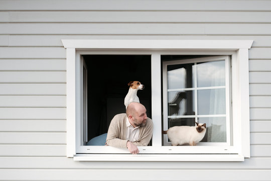 Mature man with cat and dog leaning out of window of his house