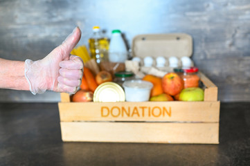 Volunteer hands in medical gloves holding donation box with various products for poor people. Pandemic coronavirus food shortages. Food donations or delivery concept. Free space for text copyspace