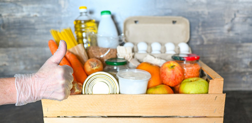 Volunteer hands in medical gloves holding donation box with various products for poor people. Pandemic coronavirus food shortages. Food donations or delivery concept. Free space for text copyspace