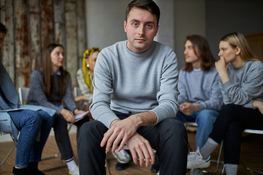 Young Addict Seriously Look At Camera, Sit Isolated In Room With Group Of People Having Conversation In The Background. Sick Male Need Help