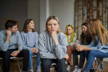 young addict seriously look at camera, sit isolated in room with group of people having...
