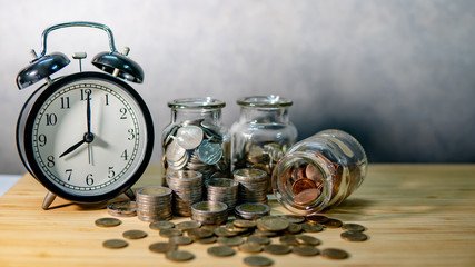 Clock with coins in currency glass jars and spilling on wooden table. Saving money for future retirement. Financial business growth. Time investment concept