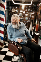 Obraz premium An elderly gray-haired and bearded man, in everyday clothes, with a smile, sits on a chair in a barbershop and waits for a specialist. Vertical frame