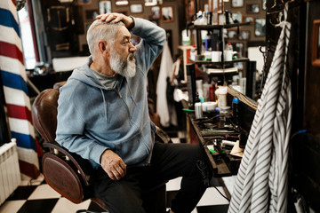 An elderly gray-haired and bearded man looking in the mirror, checks how it was cut in the new barbershop