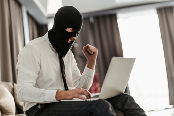 A man in a Balaclava mask and a white office shirt, sitting at a laptop and trying to anonymously...