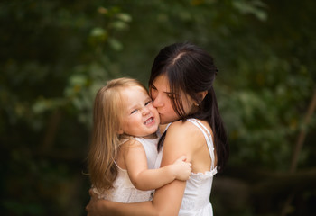 Young pretty black haired mother with blond little daughter in white dresses hugging outside in the summer