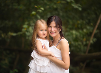 Young pretty black haired mother with blond little daughter in white dresses hugging outside in the summer