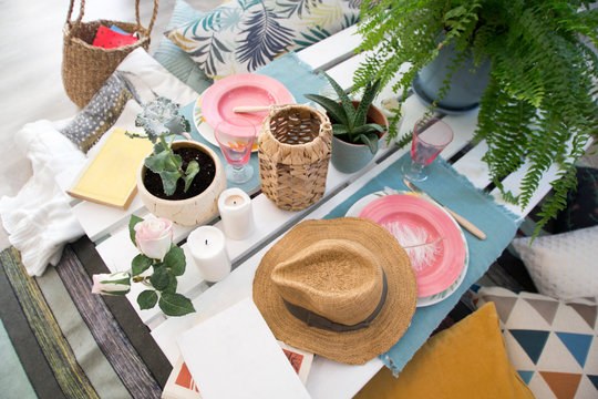 stylized outdoor Breakfast on a white hand-painted tray and a covered table with dishes, plants, candles, a book, an empty vase, pillows and a straw hat.