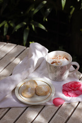 White porcelain cup of hot coffee, cappuccino with three Italian amaretti cookies.Summer breakfast in a terrace - coffee, amaretti and flowers