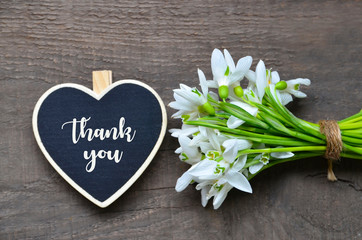 Thank You or thanks greeting card with bouquet of snowdrop flowers and decorative heart on old wooden background.International Thank You Day or Mother's Day concept. 
