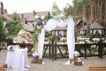 Wedding decoration. Arch with white textile curtain and purple flower decor. Wooden elements. Outdoor wedding ceremony in the coniferous forest. Selective focus