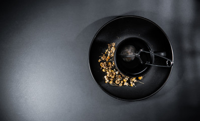 Chamomile tea cup with loose dried petals and strainer, isolated on gray black background.