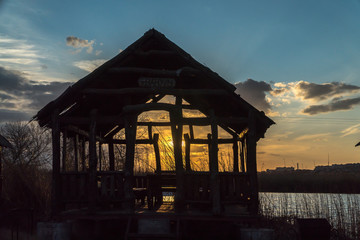 Wooden Gazebo with a thatched roof. Idyllic picnic spot, on the side of the river. Sunset time at the barbecue place. A small house at the lake.